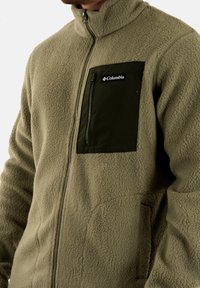 Giacca in pile verde oliva con colletto alto, tasca sul petto con zip e tasche laterali, caratterizzata da un tessuto a trama e dal logo Columbia.