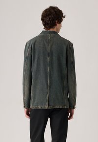 Hombre de espaldas vistiendo una chaqueta de mezclilla desvanecida con costuras visibles y pantalones negros, frente a un fondo claro y liso.