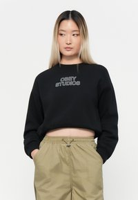 Mustad musta cropitud sviiter, millel on valge tikand "OBEY STUDIOS". Kandmiseks heleda rohelise värvi, nööriga vöökohtadega püksid, millel on küljetaskud.