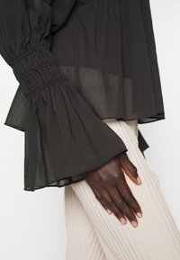 Blouse noire transparente avec manches longues évasées et froncées sur un bras à la peau foncée reposant sur un pantalon beige clair côtelé.