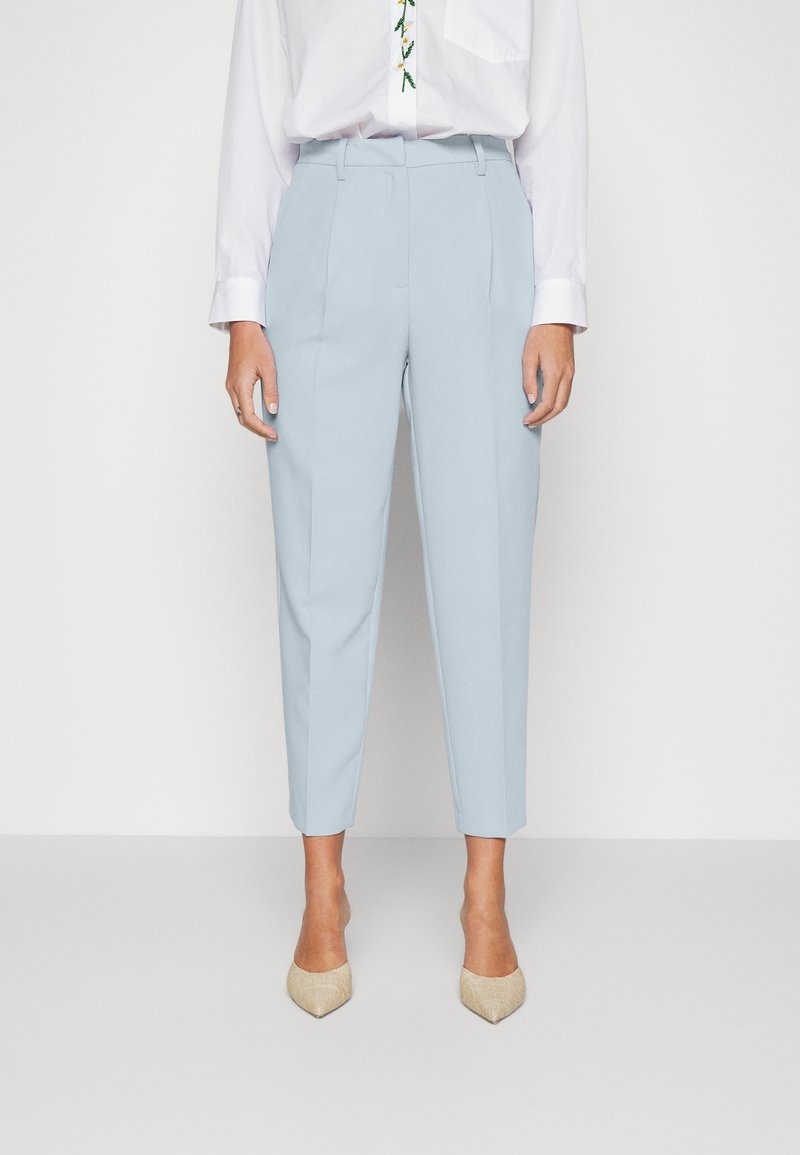 Pantalon bleu clair ajusté longueur cheville porté avec une chemise blanche à manches longues et des chaussures beiges à bouts pointus.