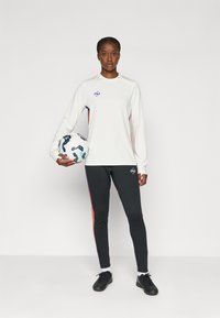 Tricou sportiv alb cu mânecă lungă și logo, purtat cu pantaloni negri cu dungi laterale roșii și mov, ținând o minge de fotbal cu un model.