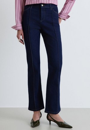Personne portant un jean droit bleu foncé, une chemise rayée rose et des chaussures pointues vert foncé à talon bas, debout sur un sol uni.