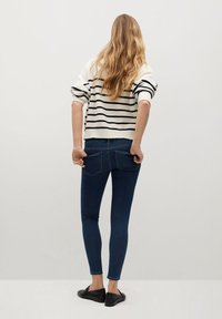 Mujer con cabello largo y ondulado, vistiendo un suéter blanco con rayas negras, jeans ajustados oscuros y mocasines negros, contra un fondo liso.