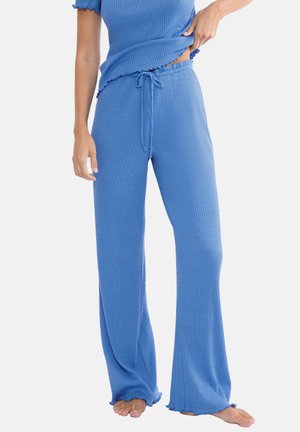 Kobieta ubrana w niebieskie, szerokie spodnie z prążkowanego dzianego materiału z sznurkiem w talii oraz dopasowany krótki top z krótkim rękawem, stojąca boso na białym tle.