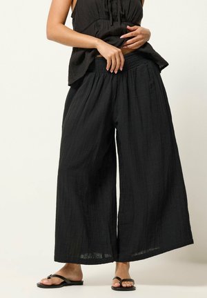 Mujer con pantalones anchos negros y sueltos con una cintura elástica alta, combinados con sandalias negras planas con tiras sobre un fondo neutro.