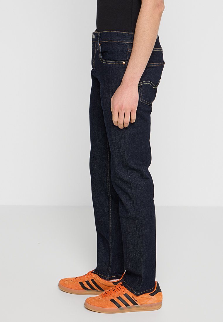 Jean en denim bleu marine avec une coupe droite, coutures visibles, cinq poches et une fermeture éclair. Associés à des baskets orange avec des rayures noires.
