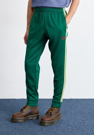 Person wearing green Adidas track pants with yellow side stripes and brown lace-up boots standing on blue carpet.
