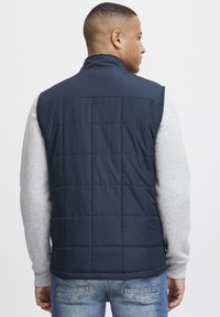 Marineblauer gesteppter Weste mit hohem Kragen, kombiniert mit einem grauen Langarmshirt. Verfügt über eine glatte Textur und ein dezentes Design.