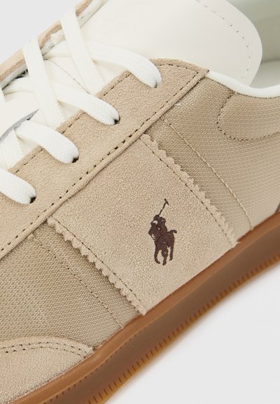 Μπεζ και ανοιχτό καφέ αθλητικό παπούτσι με λευκά κορδόνια, πάνελ από σουέτ και ύφασμα, και ένα λογότυπο παίκτη πόλο σε σκούρο καφέ στη πλευρά.