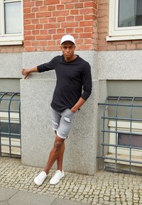 Szare rozdarcie spodenki, czarna koszula z długim rękawem, biała czapka i białe trampki. Osoba opiera się o doskonale teksturowaną kamienną ścianę.