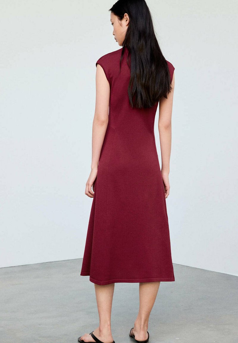 Femme aux longs cheveux noirs portant une robe midi bordeaux sans manches et des sandales noires, debout sur un sol gris contre un mur blanc.