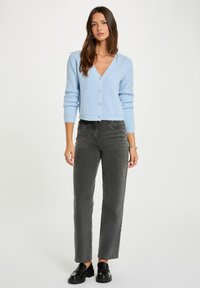 Hellblauer gestrickter Cardigan mit V-Ausschnitt, fünf dekorativen Knöpfen, kombiniert mit hochgeschnittenen, geraden dunkelgrauen Jeans und schwarzen Schuhen.