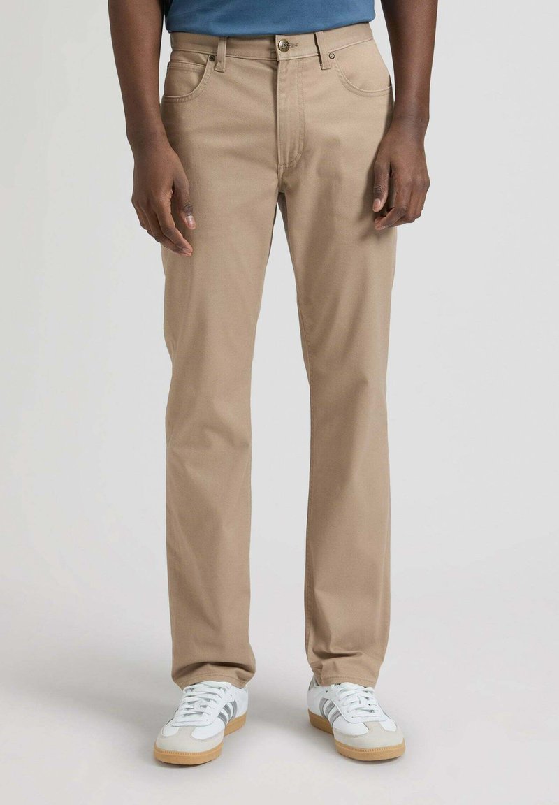 Hombre con pantalones beige de pierna recta, zapatillas blancas con rayas grises y camisa azul, de pie contra un fondo liso.