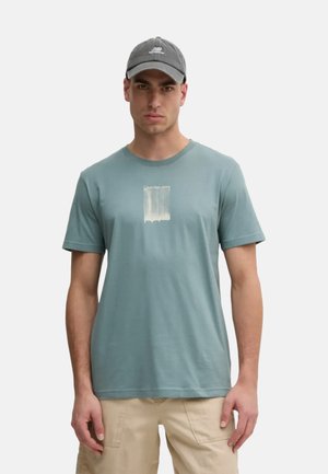 Camiseta de algodón azul claro con mangas cortas, con un estampado gráfico en blanco; combinada con una gorra gris. Ajuste casual y cuello redondo.