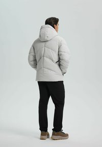 Hellgraue Daunenjacke mit Kapuze, ausgestattet mit einem geometrischen Steppmuster, kombiniert mit schwarzer Hose und braunen Wanderschuhen.