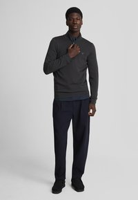 Pullover grigio con zip e finitura testurizzata, indossato sopra una maglietta scura. Abbinato a pantaloni blu navy e scarpe nere, evidenziando uno stile casual.