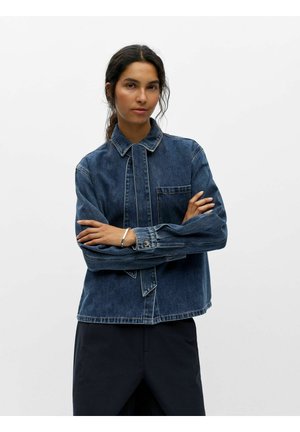 Femme aux cheveux foncés, debout les bras croisés, portant une veste en jean bleue et un pantalon noir sur un fond blanc uni.