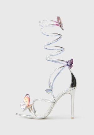 Sandalia plateada de tacón alto con tira alrededor del tobillo en espiral decorada con coloridos adornos de mariposa sobre un fondo blanco.