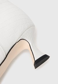 Zapato de tacón alto blanco con patron de cocodrilo texturizado, suela beige y punta negra en el tacón, sobre un fondo blanco.