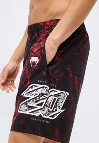 Άνδρας που φοράει μαύρο και κόκκινο σορτς με φερμουάρ στο τσέπη και το γραφικό "20ή Επέτειος 2005-2025" στο πλάι.