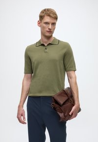 Polo manches courtes vert olive en tissu texturé, avec bordure de col noire, associé à un pantalon bleu marine et tenant un sac messager en cuir marron.