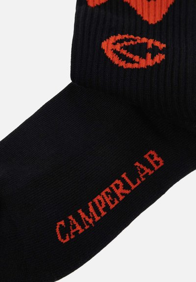 Chaussette noire avec texte "CAMPERLAB" rouge-orange sur le pied et un motif graphique rouge-orange partiellement visible sur la bordure côtelée.