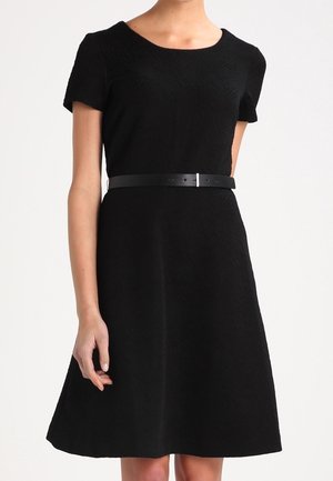 Femme portant une robe noire courte à manches courtes, coupe évasée, avec un col rond et une fine ceinture noire à la taille.
