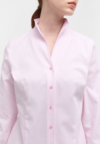 Blusa rosa chiaro con un motivo testurizzato, colletto alto e bottoni rosa. Il tessuto appare liscio e tagliato per un look aderente.