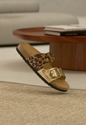 Σανδάλι slide με δύο ρυθμιζόμενους ιμάντες με πόρπες, ένας με λεοπάρ σχέδιο και ένας μεταλλικός χρυσός, που βρίσκεται σε μπεζ χαλί κοντά σε ξύλινο τραπέζι.