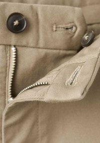 I pantaloni beige presentano una chiusura con cerniera metallica, un grande bottone di plastica e un'etichetta di stoffa cucita in vita.