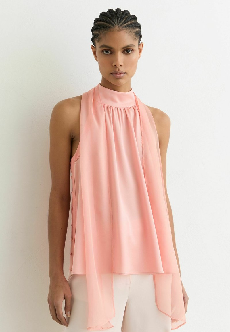 Fiatal nő fonott hajjal, világos rózsaszín, ujjatlan, magas nyakú blúzban, amelynek sál részletei lebegnek, fehér nadrággal.