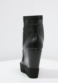 Botín de tacón cuña negro con un exterior de cuero suave, paneles laterales elásticos y una suela de goma texturizada. Diseño elegante con punta redondeada.