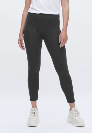 Schwarze, hochtaillierte Leggings aus dehnbarem Stoff mit glatter Textur und figurbetontem Design. Kombiniert mit weißen Sportschuhen.