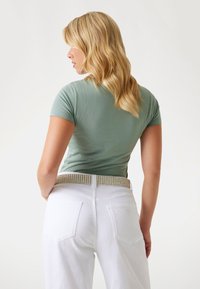 T-shirt fitted verde menta con maniche corte, abbinato a jeans bianchi a vita alta. Accentua la cintura con borchie e i capelli texturizzati sono visibili.