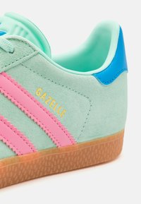 Mintgrønn semsket sneaker med rosa tre-stripes design og gyllent "GAZELLE"-bokstaver. Har en blå hæl-akseent og gummi såle tekstur.