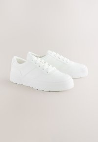 Baltos sportbačiai su lygiu, tekstūruotu viršutiniu sluoksniu, pakilusia gumine pada, apvalia nosimi ir užsegimu antvarstais su šešiais skylais.