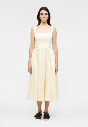 Γυναίκα που στέκεται με το πρόσωπο προς τα εμπρός, φοράει ένα αμάνικο μπεζ μίντι φόρεμα με τετράγωνη λαιμόκοψη και καφέ δερμάτινα παπούτσια με κορδόνια.