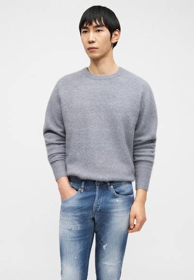 Άνδρας που φοράει ανοιχτό γκρι χνουδωτό πουλόβερ με στρογγυλή λαιμόκοψη και ξεθωριασμένο μπλε τζιν με το χέρι του στην αριστερή τσέπη, στέκεται μπροστά σε λευκό φόντο.
