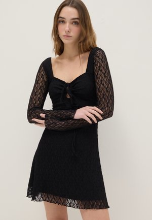 Giovane donna con capelli lunghi che indossa un abito nero in pizzo con maniche lunghe e un dettaglio con fiocco frontale, in piedi davanti a uno sfondo chiaro uniforme.