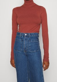 Turtleneck trui in roest, gemaakt van zachte stof. Gecombineerd met hooggesneden blauwe spijkerbroek, met twee voorzakken en een knoopsluiting.