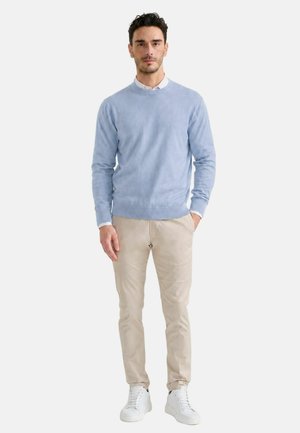 Uomo con maglione azzurro chiaro, camicia bianca, pantaloni beige e sneakers bianche, in piedi con una mano in tasca su sfondo neutro.