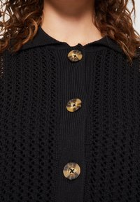 Svart stickad cardigan med en texturerad design, stora runda knappar i ljusbrun och mörkbrun, samt en krage vid halsringningen.