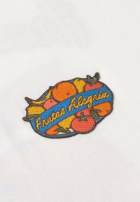 Patch brodé coloré sur tissu blanc montrant des fruits variés et le texte "Frutas Alegria" sur une bannière bleue.