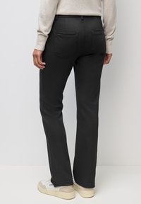 Persona che indossa pantaloni neri a gamba dritta, maglione beige e scarpe da ginnastica bianche, in piedi contro uno sfondo chiaro e uniforme.