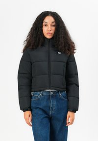 Black cropped puffer jacket with a high collar, featuring zip closure, horizontal stitching, and a small logo patch on the chest.