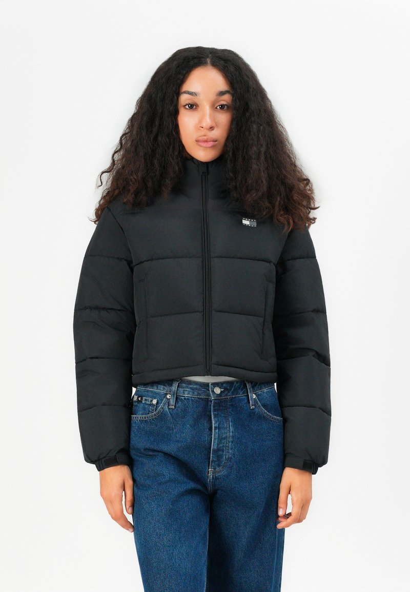 Black cropped puffer jacket with a high collar, featuring zip closure, horizontal stitching, and a small logo patch on the chest.