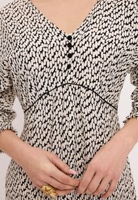 Czarna i biała sukienka w wzorki z dekoltem w kształcie litery V, trzema ozdobnymi guzikami, marszczonym pasem i falbankami przy rękawach. Gładka tekstura materiału.