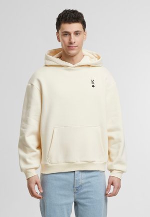 Cream hooded sweatshirt with a front pocket and ribbed cuffs, featuring a small black logo on the left chest. Soft, textured fabric.