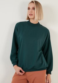 Sweatshirt verde scuro a maniche lunghe con un collo alto, polsini e orlo a costine. Texture liscia, vestibilità rilassata, abbinato a pantaloni marrone chiaro.